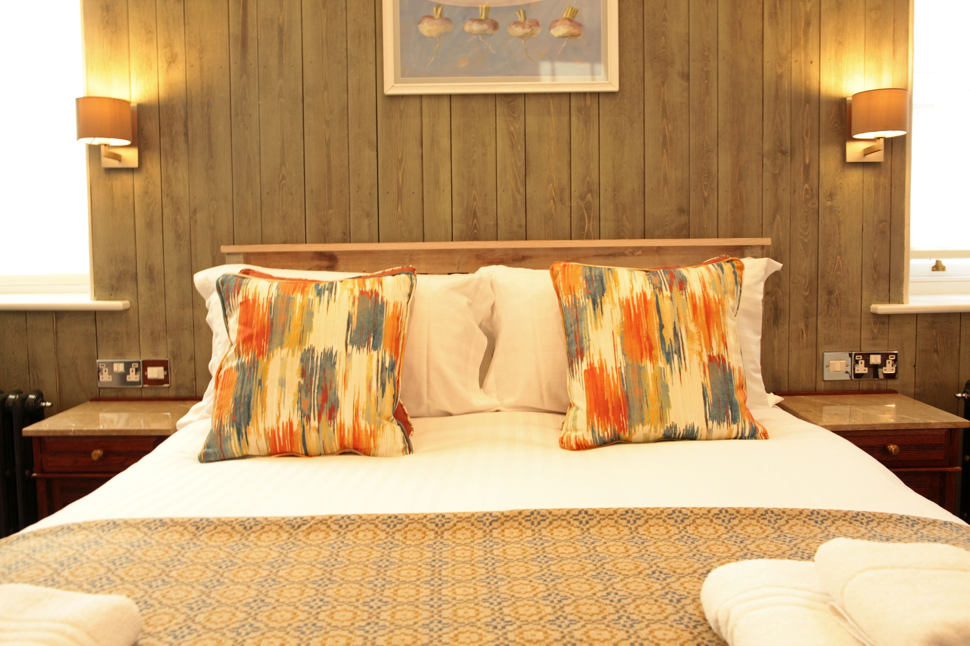 Una cama bien hecha con ropa de cama estampada, paredes de paneles de madera y dos lámparas fijadas a la pared.