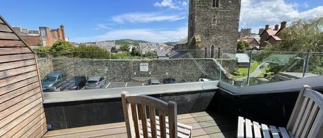 Una terraza de madera con dos sillas, un muro de piedra y una torre al fondo.