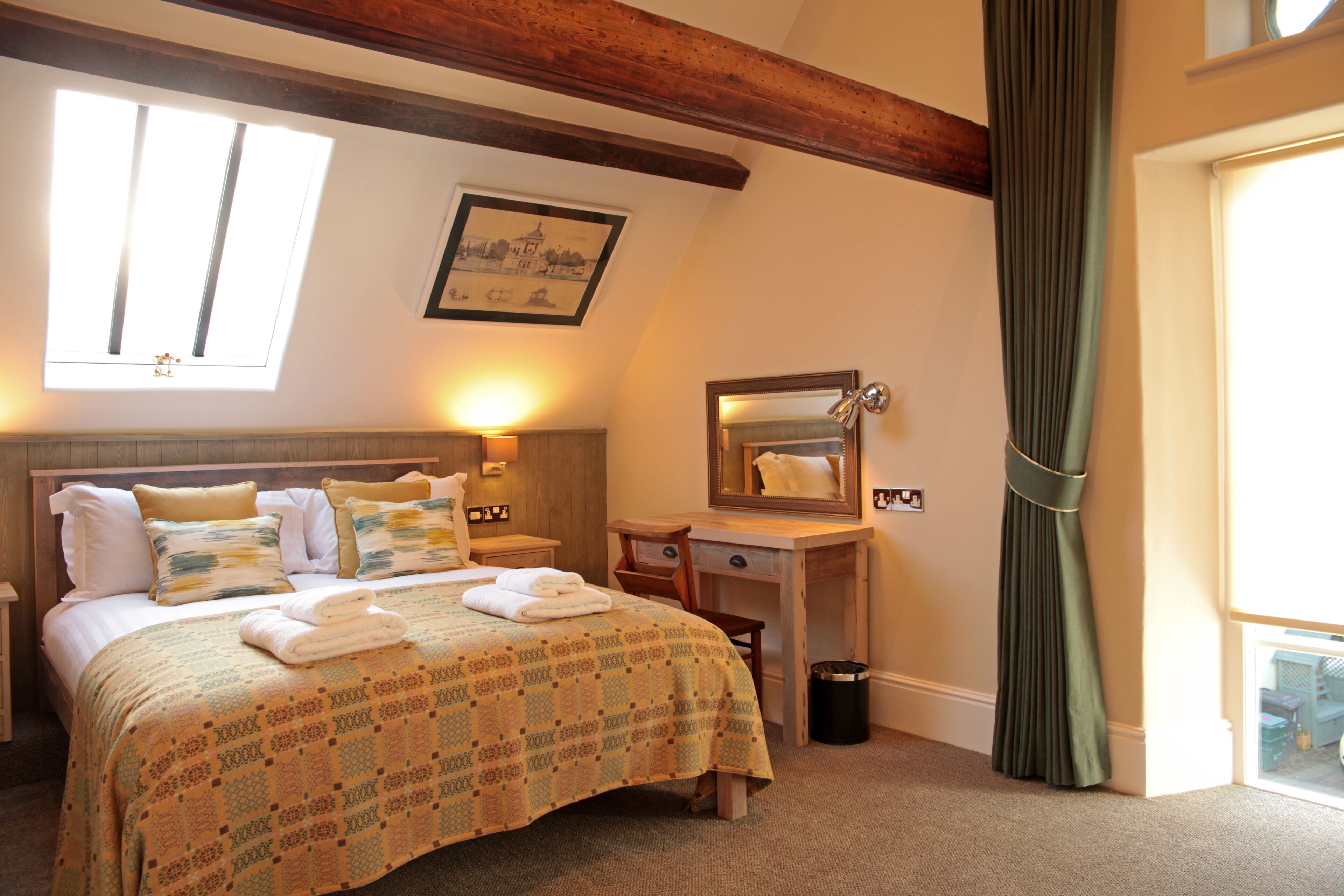 A hotel room with a bed, a desk, a mirror, a window with curtains, and a picture on the wall.