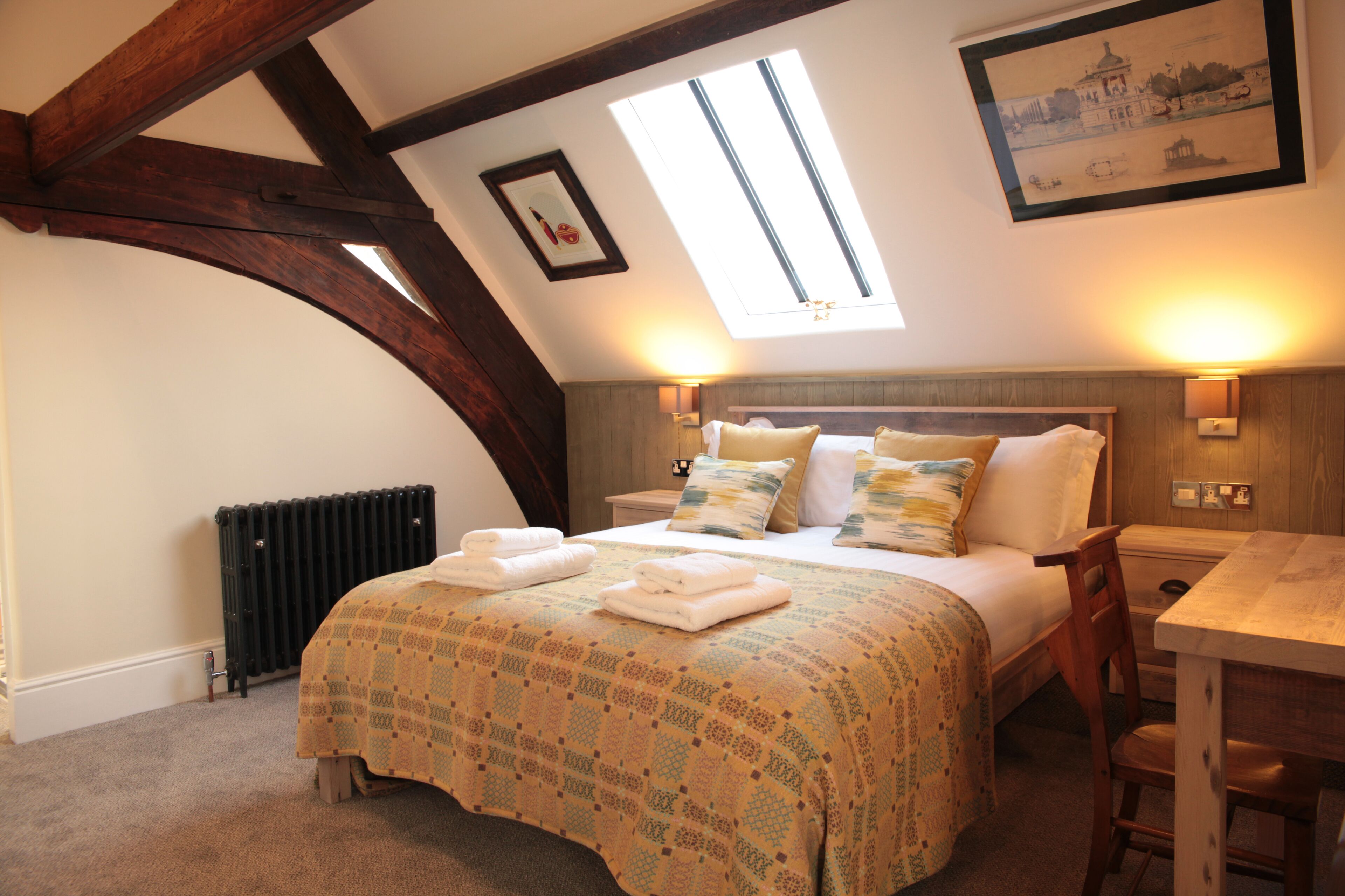 A bedroom with a sloped ceiling, a bed with a patterned bedspread, a wooden chair, and a table.