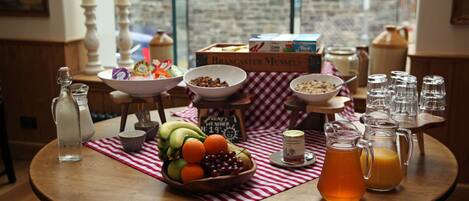 Una mesa de desayuno con un bol de fruta, cereales y una jarra de jugo de naranja.
