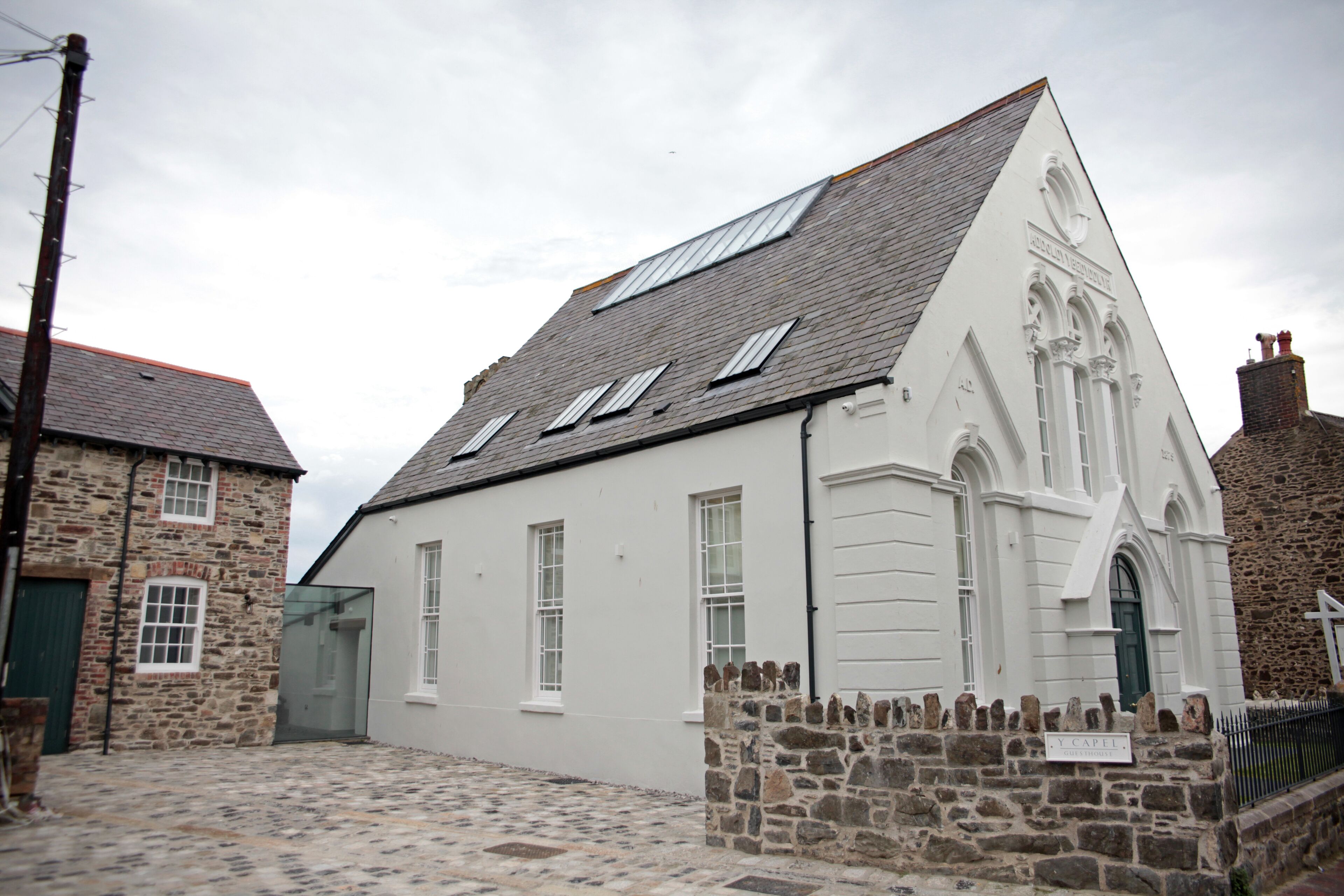 A white building with a stone foundation, a sloped roof, and a sign reading "L. Capel."