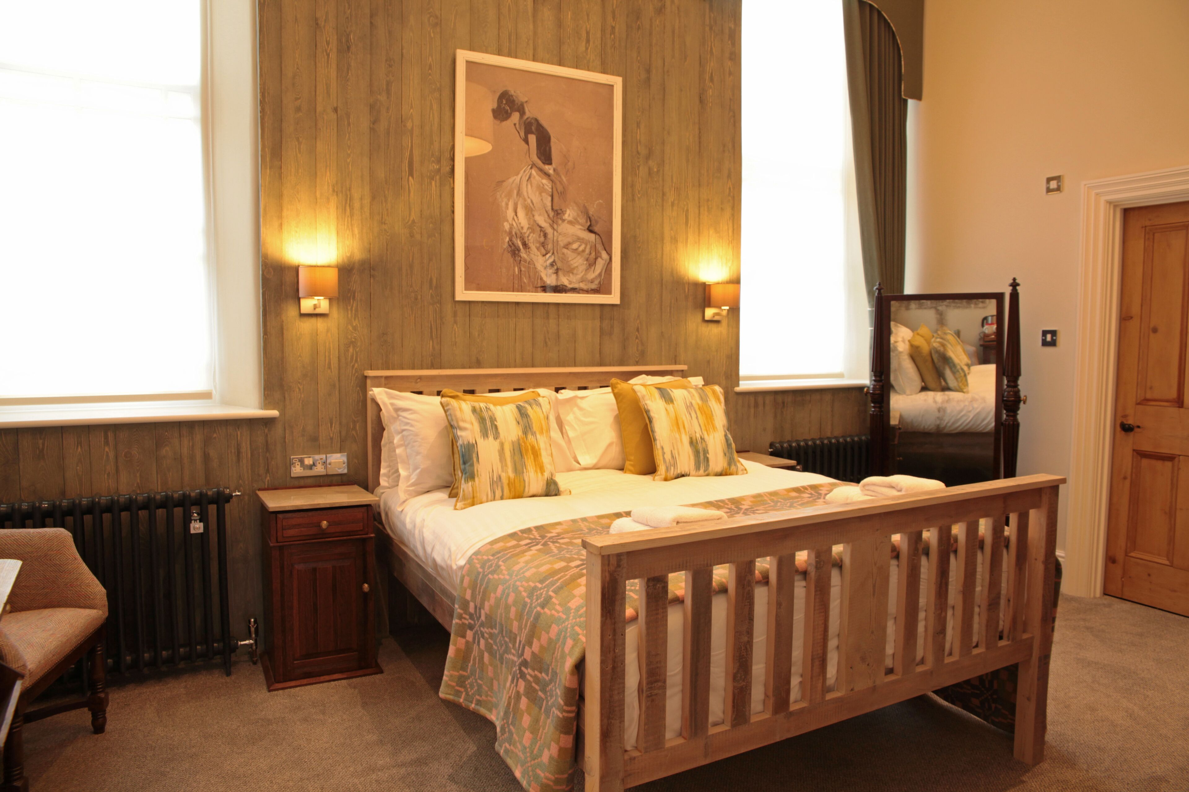 A bedroom with a wooden bed, two bedside lamps, a nightstand, a chair, a radiator, and a painting on the wall.