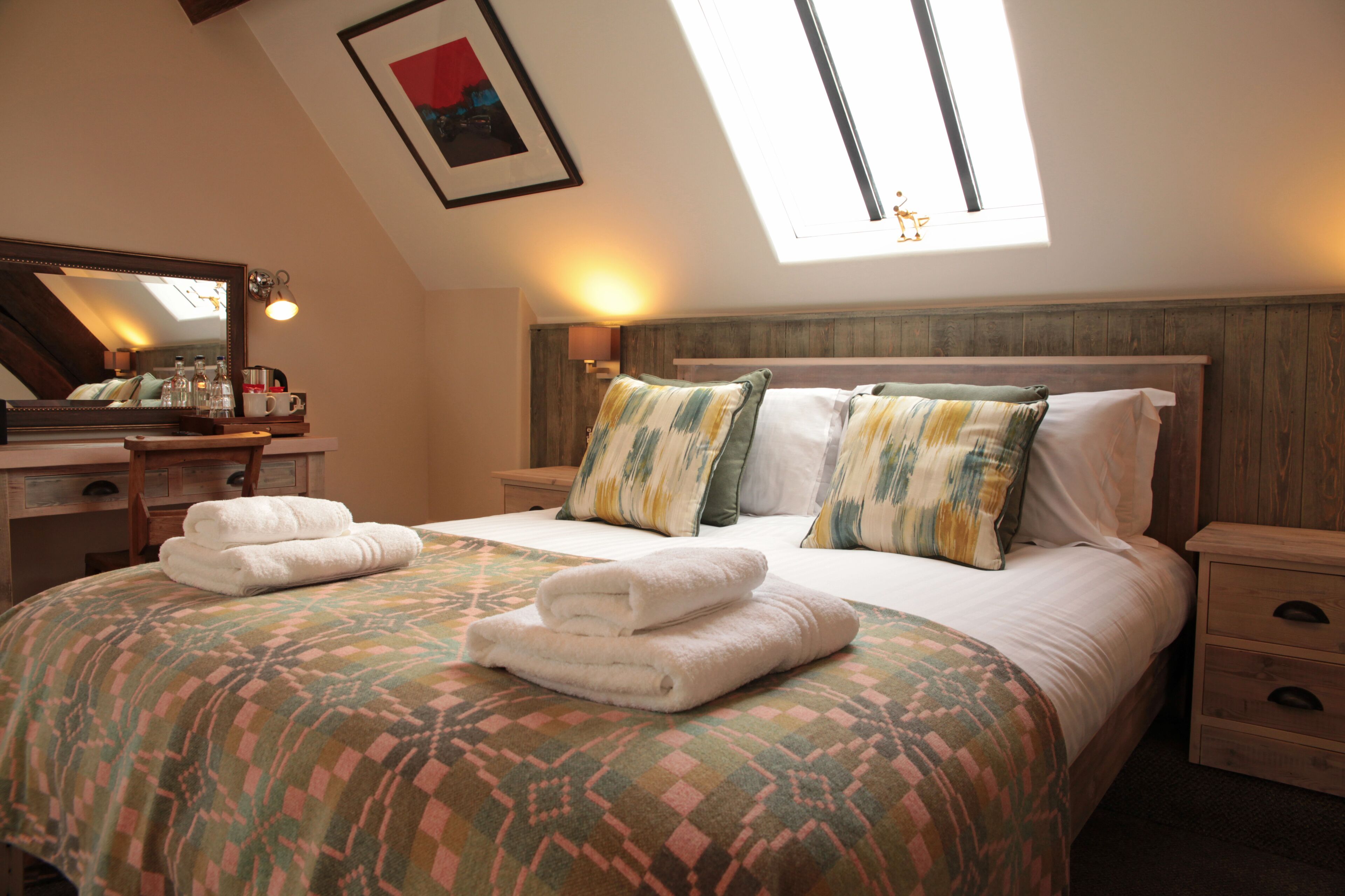 A neatly made bed with a patterned bedspread, two pillows, and two folded towels on the side. There is a nightstand with a lamp and a mirror above it.