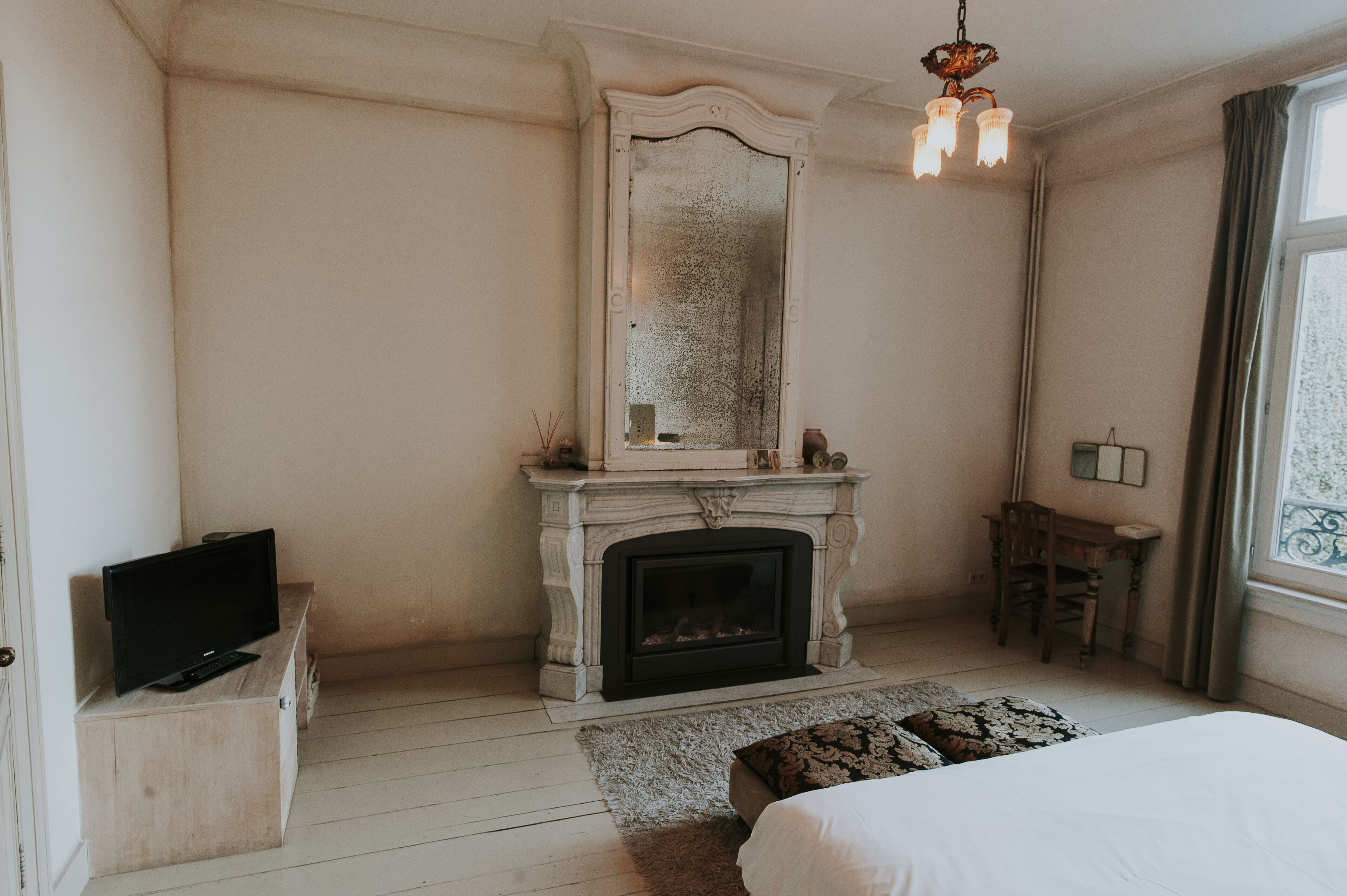 A bedroom with a fireplace, a television on a wooden dresser, a bed, and a window with curtains.
