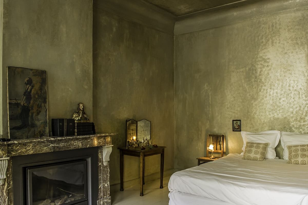 A bedroom with a bed, a nightstand, a mirror, a fireplace, and a painting on the wall.