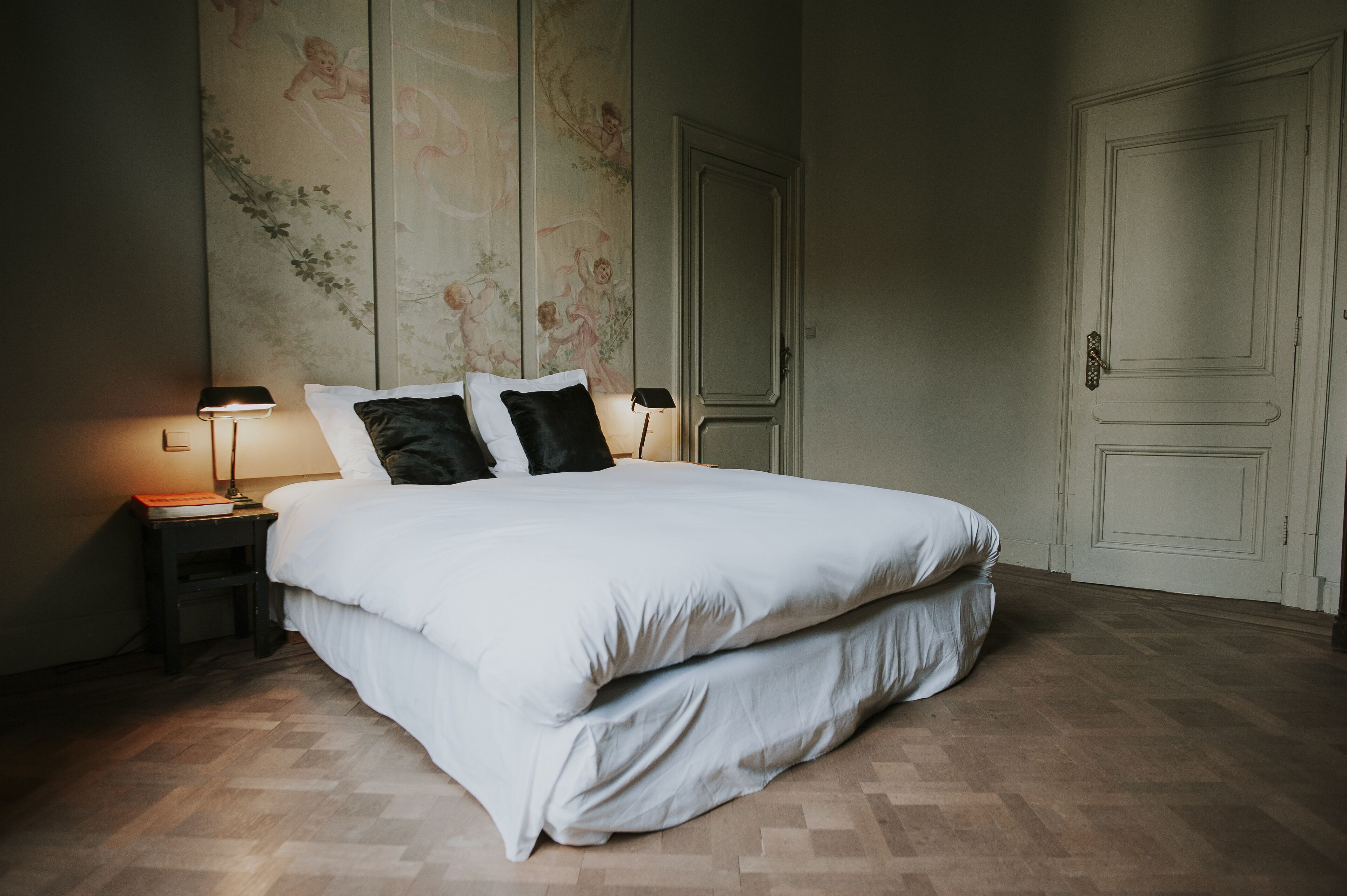 A neatly made bed with white bedding and two black pillows, flanked by two bedside tables with lamps, in a room with wooden flooring and a door in the background.