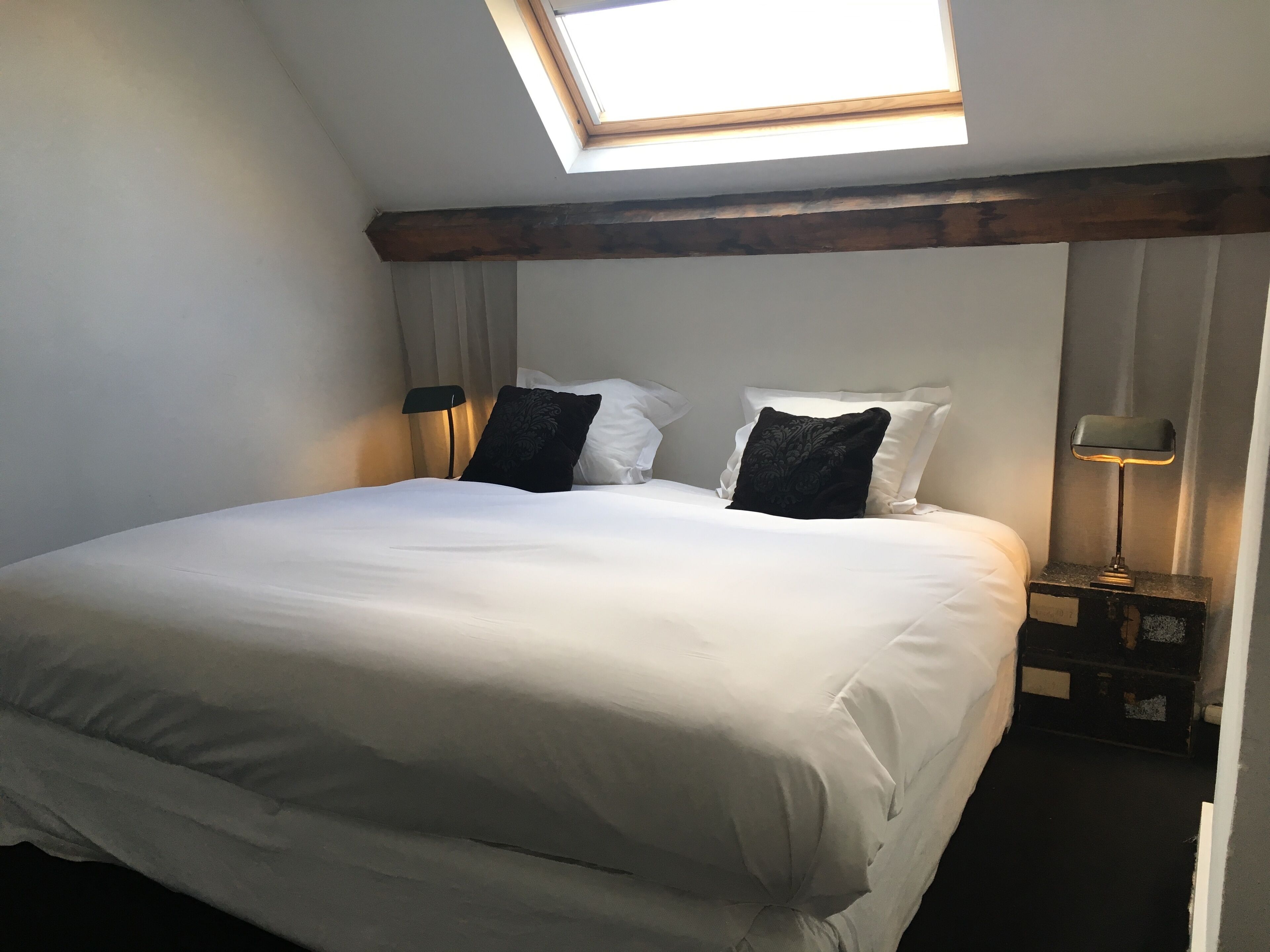 A neatly made bed with white linens, a skylight above, and a nightstand with a lamp.