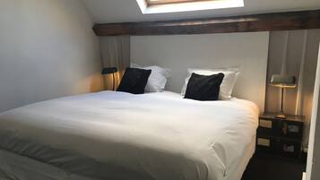 A neatly made bed with white linens, a skylight above, and a nightstand with a lamp.