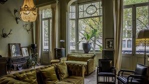 A cozy living room with a green sofa, a black armchair, and a coffee table. There are large windows with curtains, a chandelier, and wall-mounted deer antlers. - Boulevard Leopold Rooms & Suites (Antwerp)