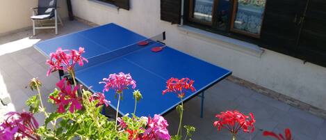 A blue ping pong table in an outdoor area with potted plants and a chair.