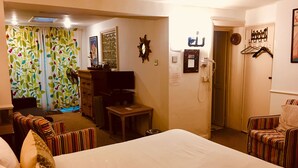 Habitación de hotel con una cama grande, televisión, escritorio y silla. Se ve una ventana con cortinas y una puerta que da a otra habitación.