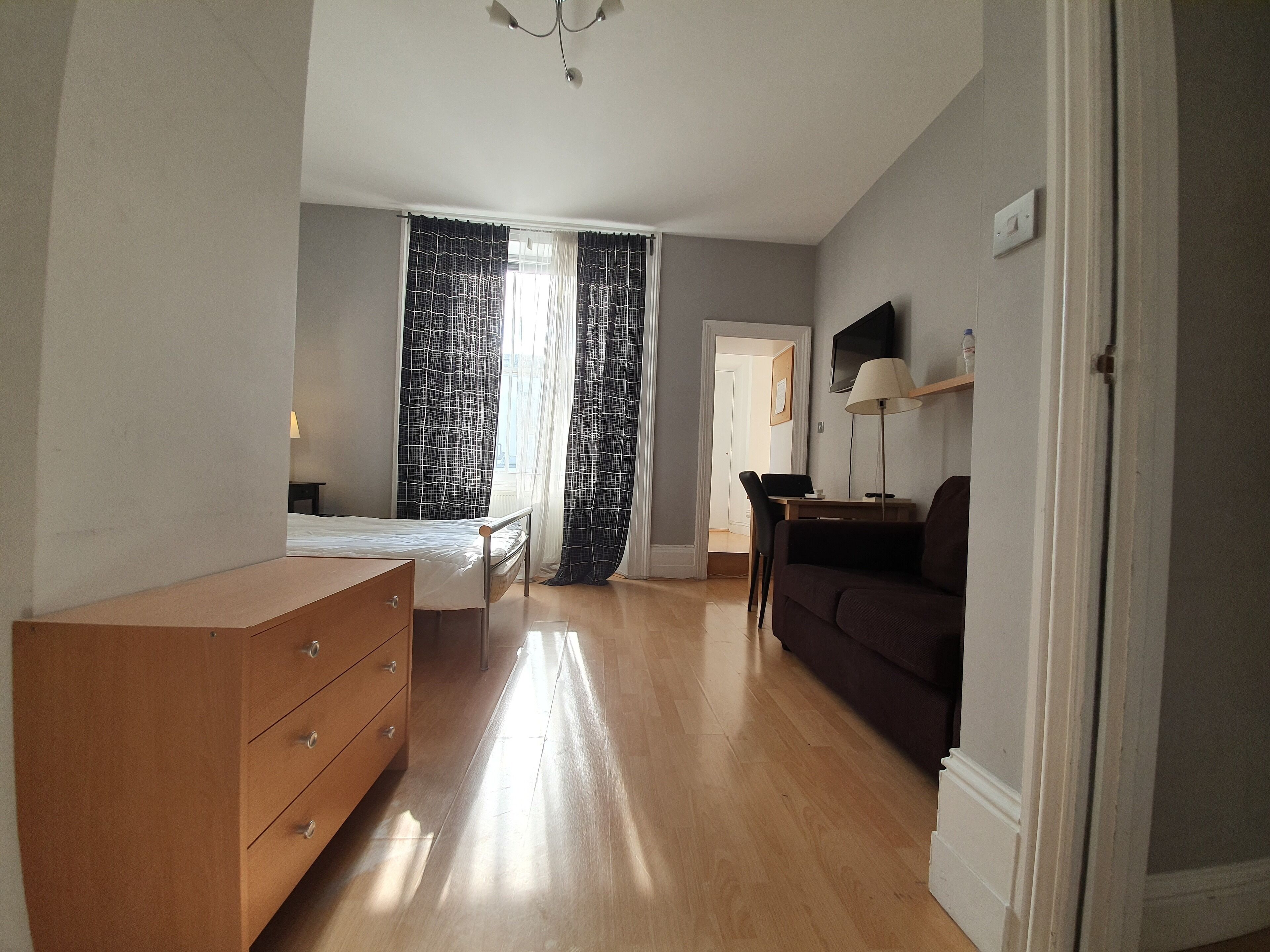 A room with a bed, a wooden dresser, a desk with a chair, a dark sofa, and a TV mounted on the wall.