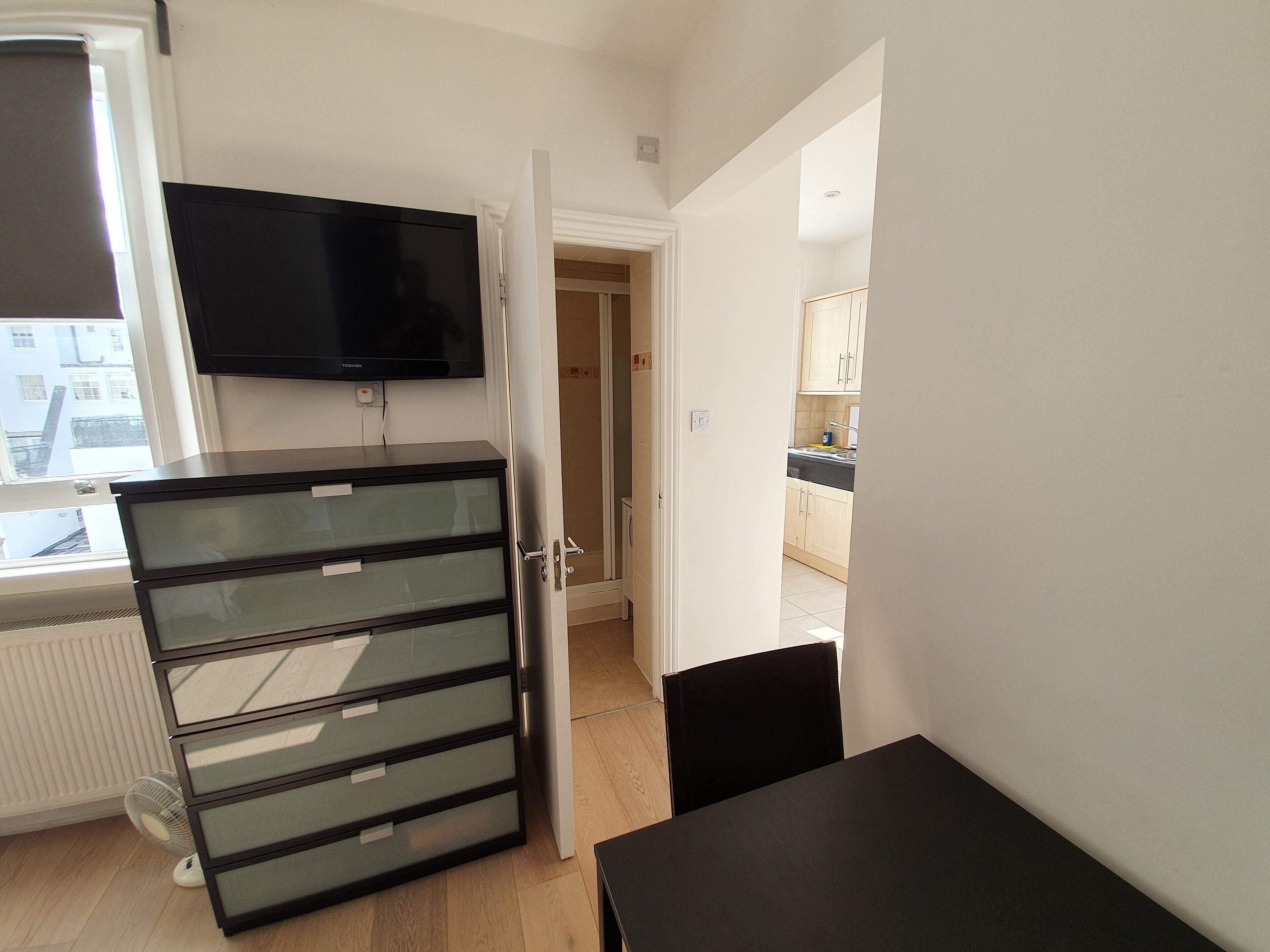 A compact living space with a kitchenette, a flat-screen TV mounted above a black filing cabinet, and a dining area with a table and chairs.