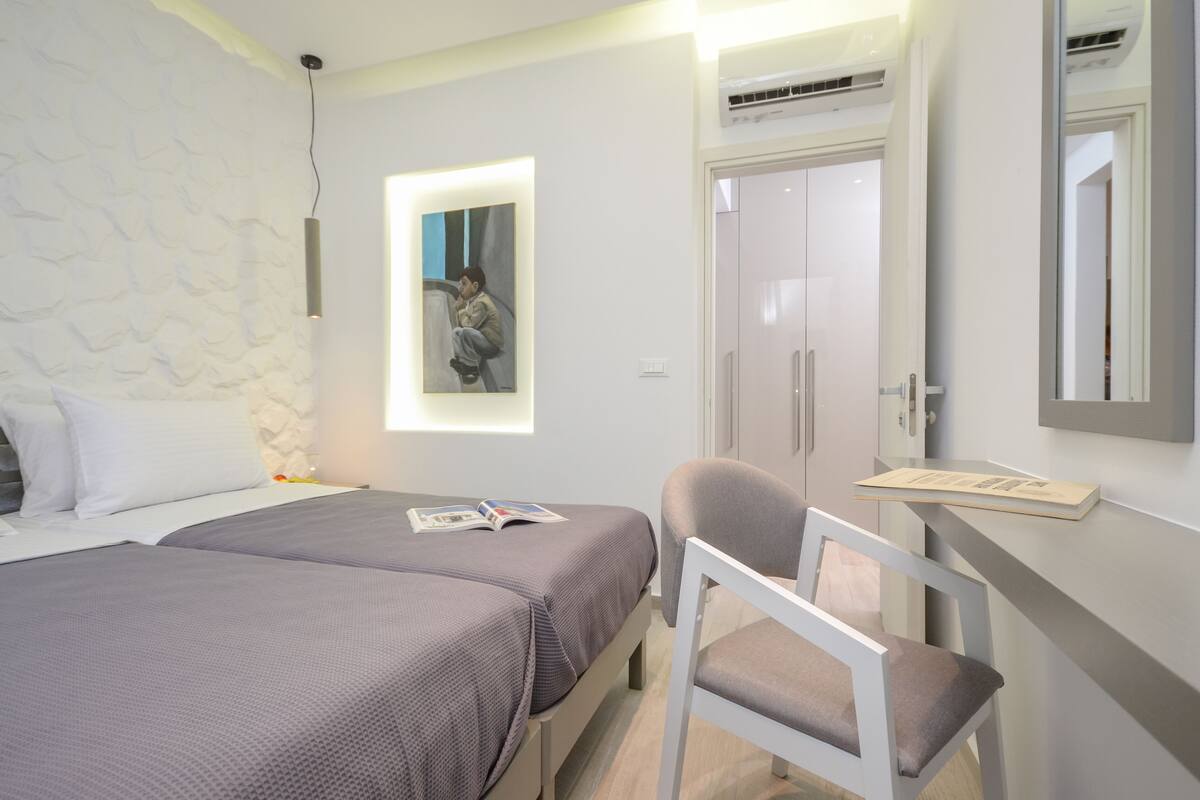 Un dormitorio con dos camas, un escritorio con silla, un armario y una ventana con un cuadro.