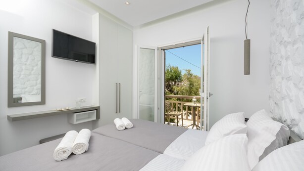 A modern hotel room with a large bed, a TV mounted on the wall, and a balcony with a table and chairs. - Elegant Apartments (Naxos)