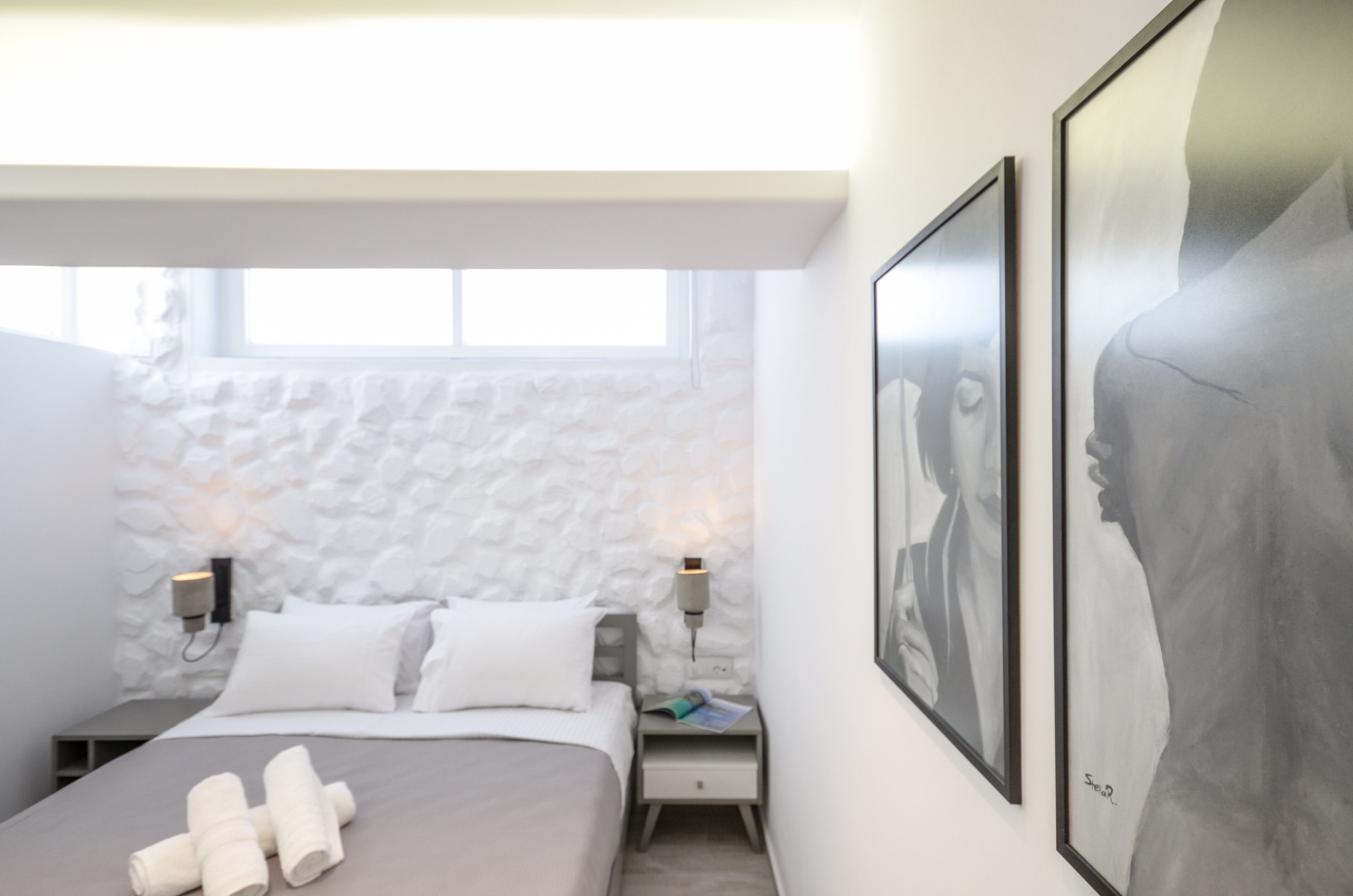 Ein modernes Schlafzimmer mit Steinwand, einem Bett mit weißen Leinentextilien, zwei Nachttischen mit Lampen und gerahmten Kunstwerken an der Wand.