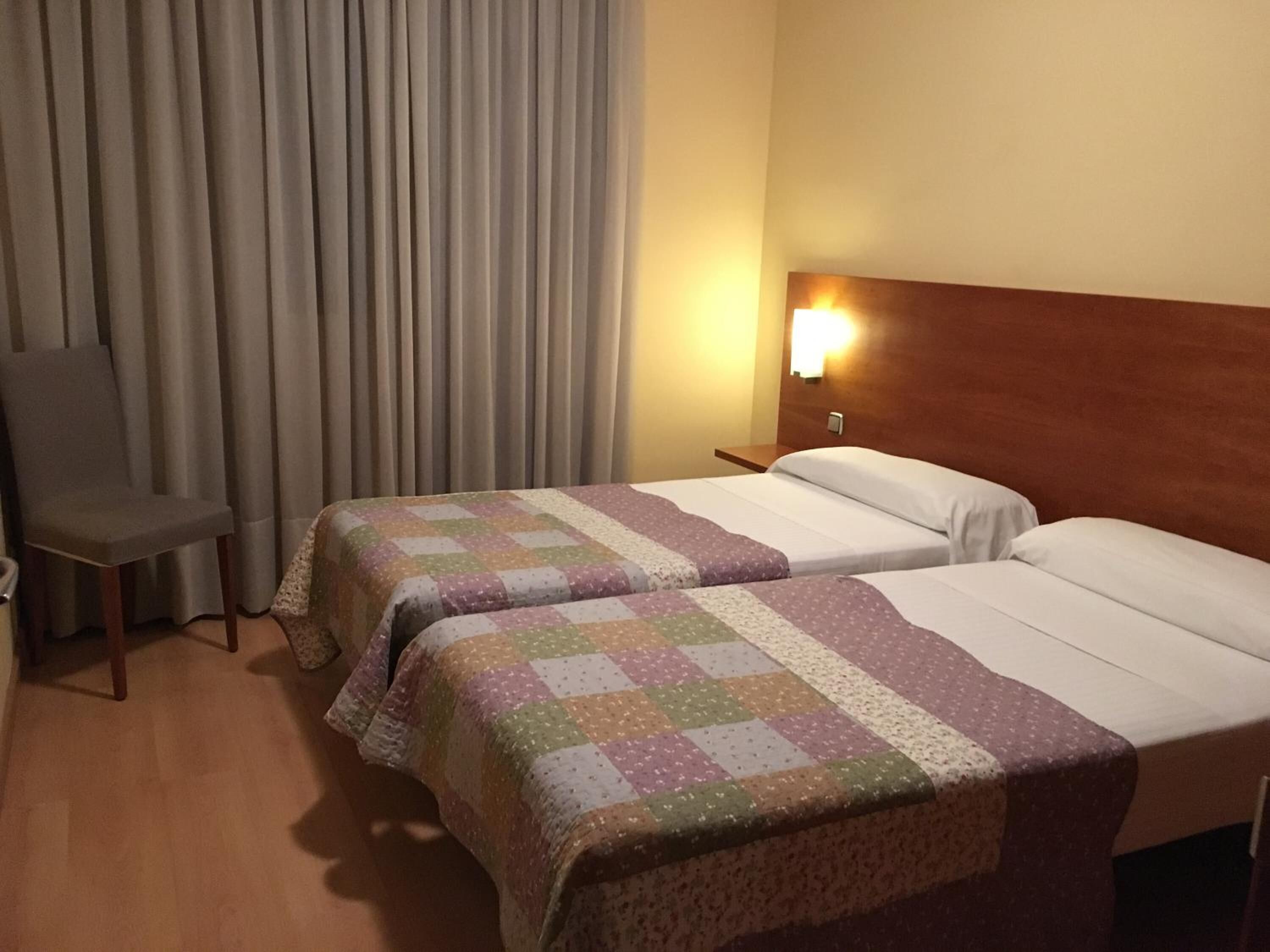 A hotel room with two beds, a chair, a curtain, and a wall-mounted light.