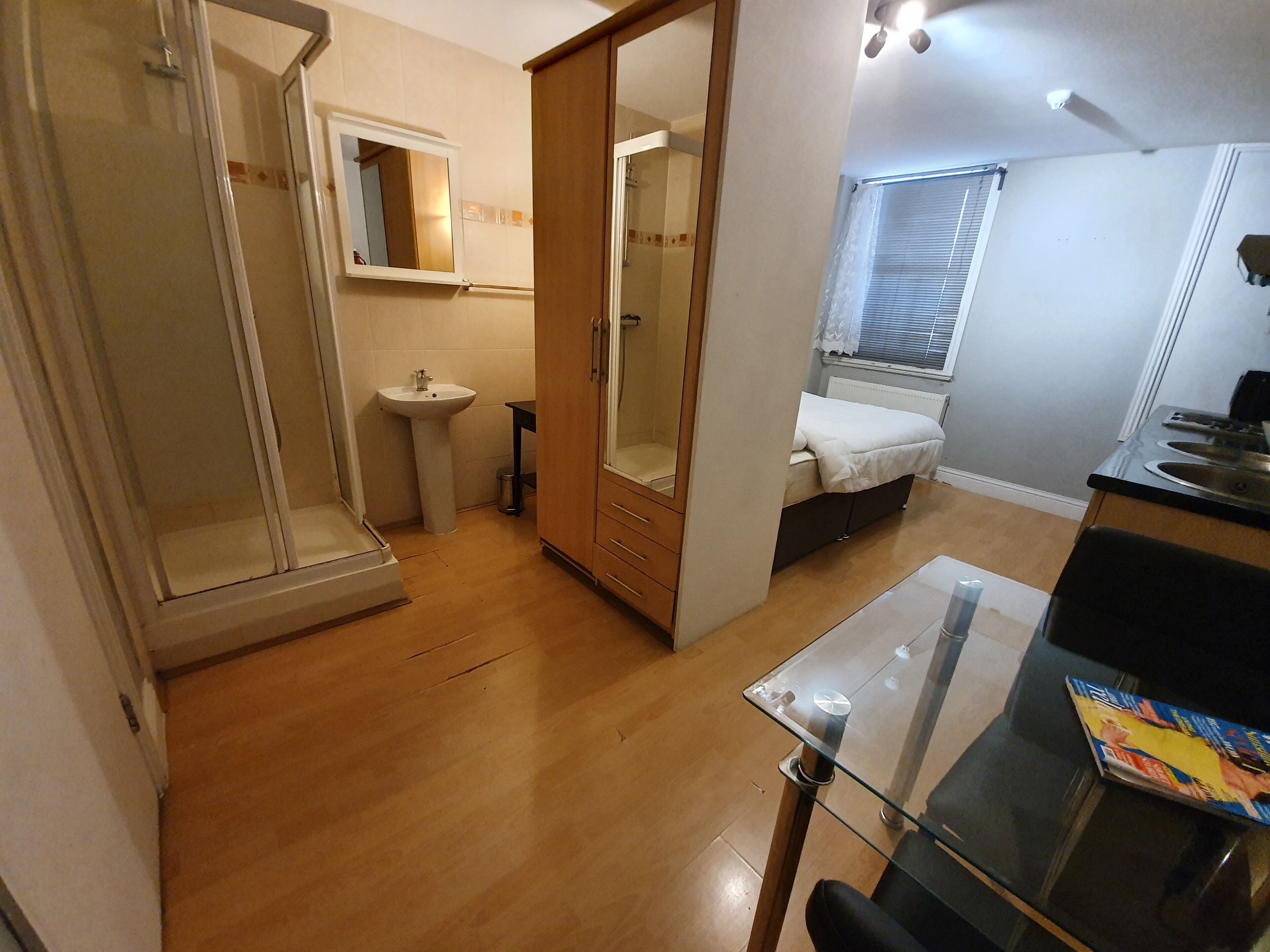 A compact bedroom with a glass table, a bedside table, a wardrobe, a mirror, a small sink, and a window with blinds.