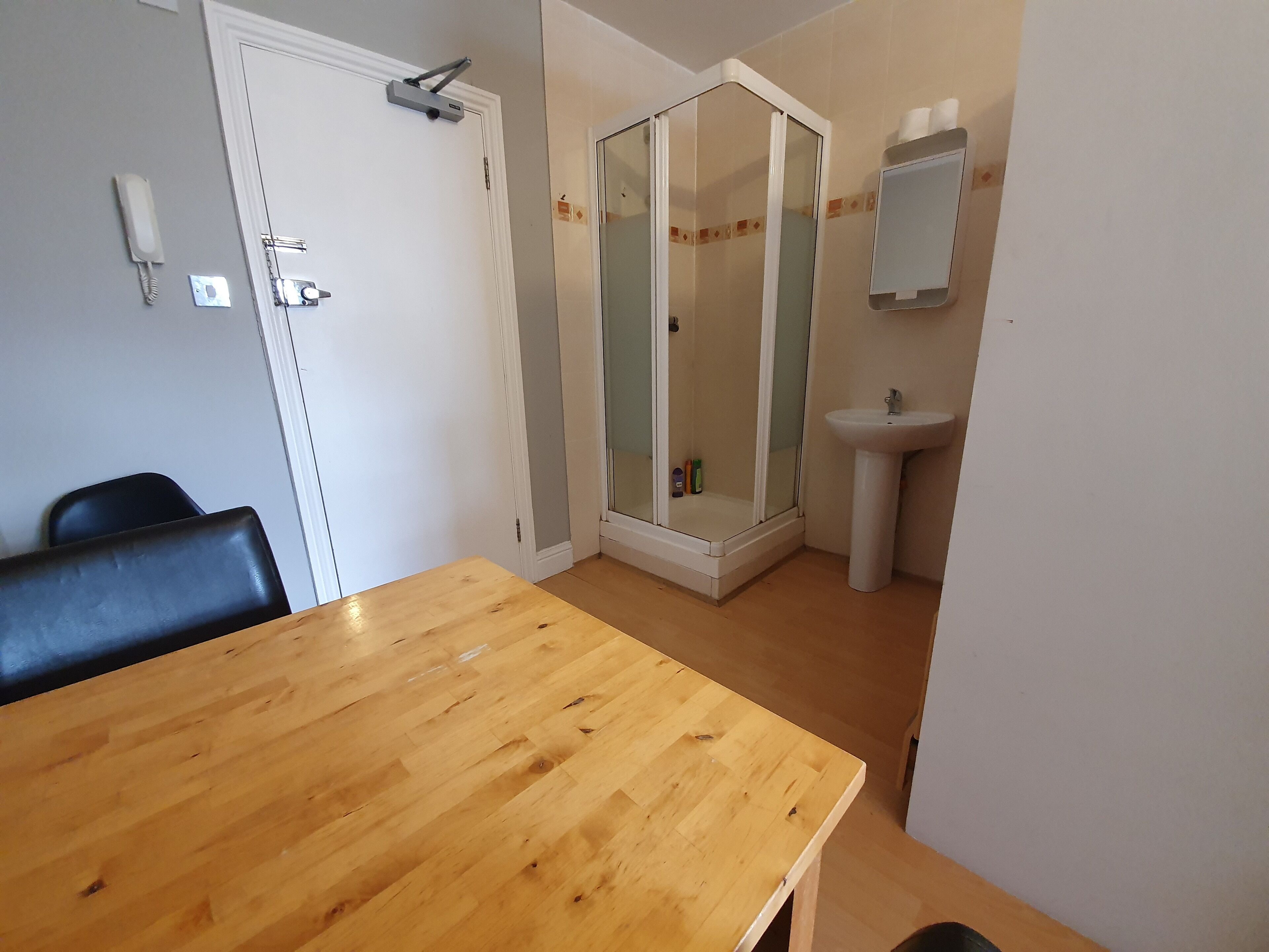 Una habitación con una mesa de madera, una silla negra, una puerta blanca, un box de ducha de vidrio y un lavamanos blanco.