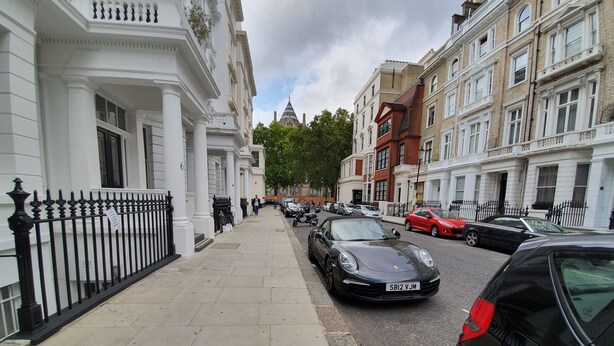 A street view with parked cars, a pedestrian, and buildings with classical architecture. - Studio Apartment in South Kensington 16 (London)