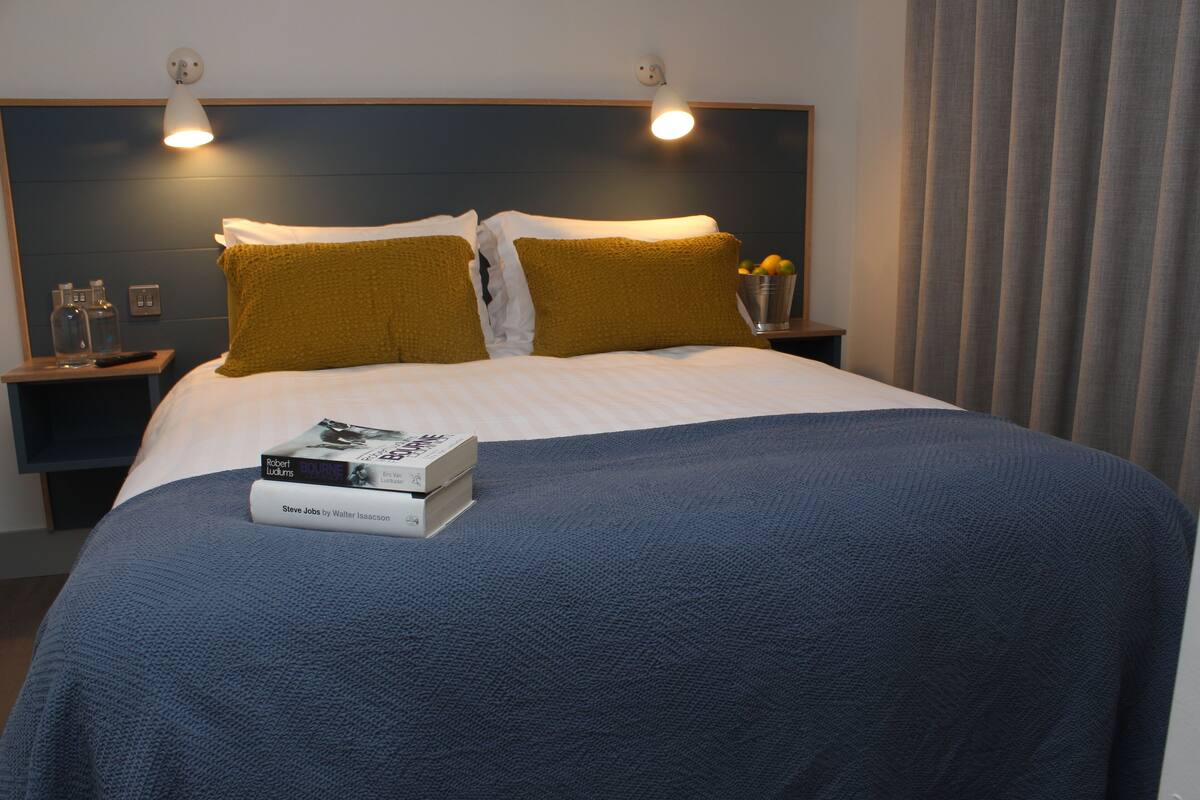 A neatly made bed with a blue blanket and two mustard pillows, a bedside table with a remote control, and two books titled 'Robert Ludlum' and 'Steve Jobs by Walter Isaacson'.