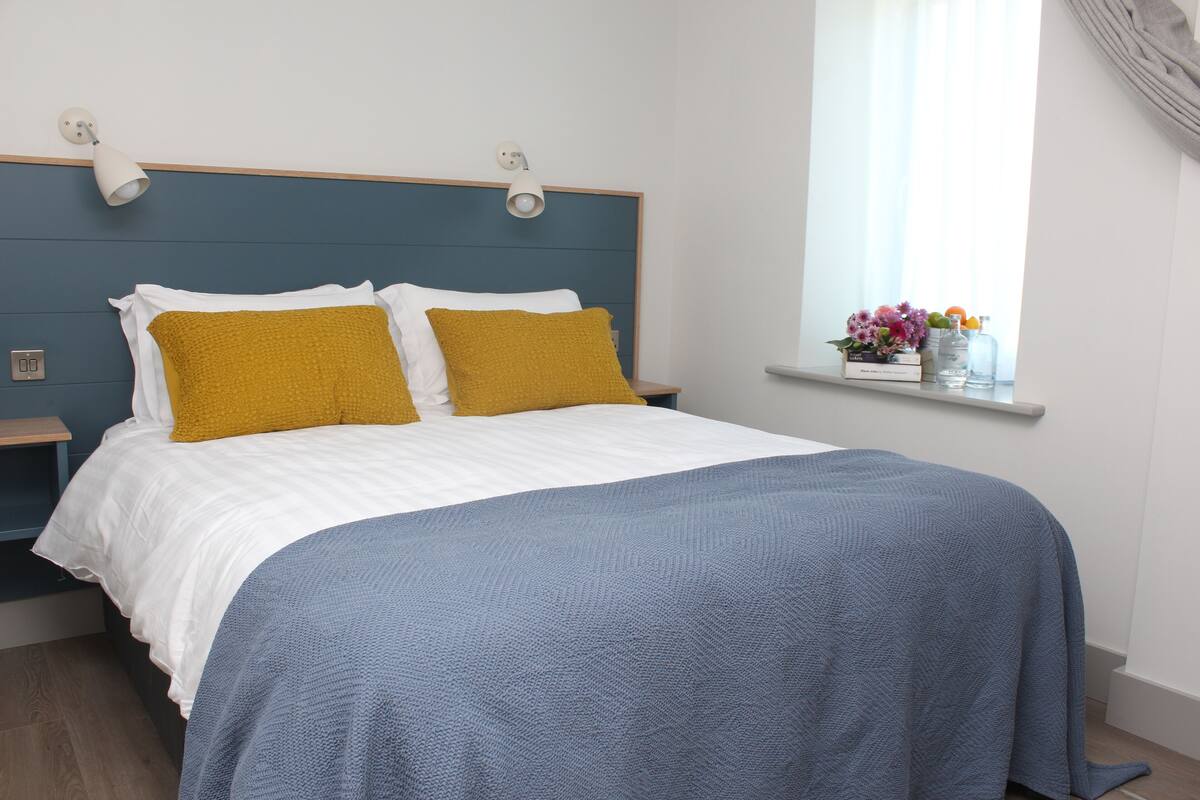 A neatly made bed with white and mustard pillows, a blue blanket, and a bedside table with a lamp.