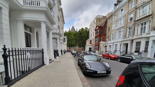 A street view with parked cars, a pedestrian, and buildings with classical architecture.