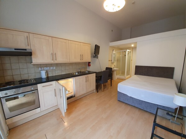 A compact hotel room with a kitchenette, a bed, a bedside table, and a shower area.