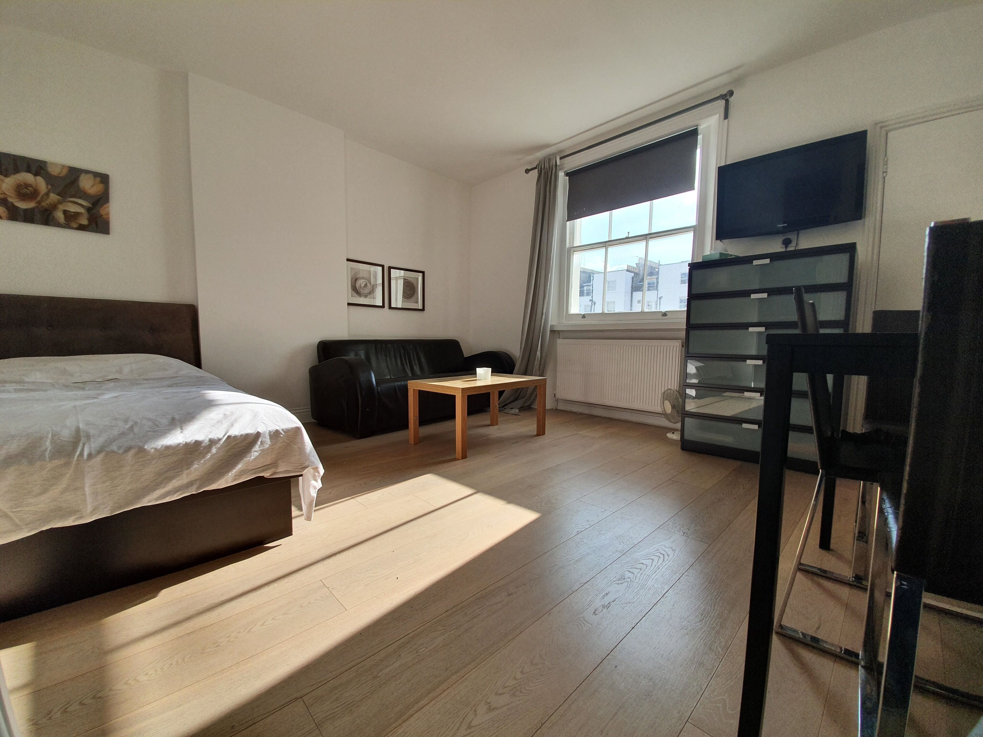 Een hotelkamer met een bed, een houten tafel, een zwarte leren bank, een televisie en een raam met gordijnen.