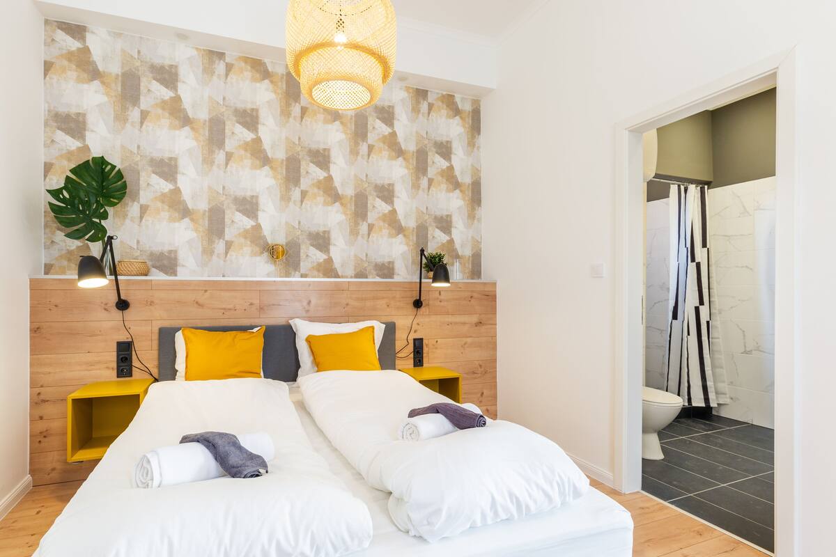 A modern hotel room with a wooden headboard, a large bed with white linens, two bedside tables with lamps, and a geometric-patterned wall above the bed.