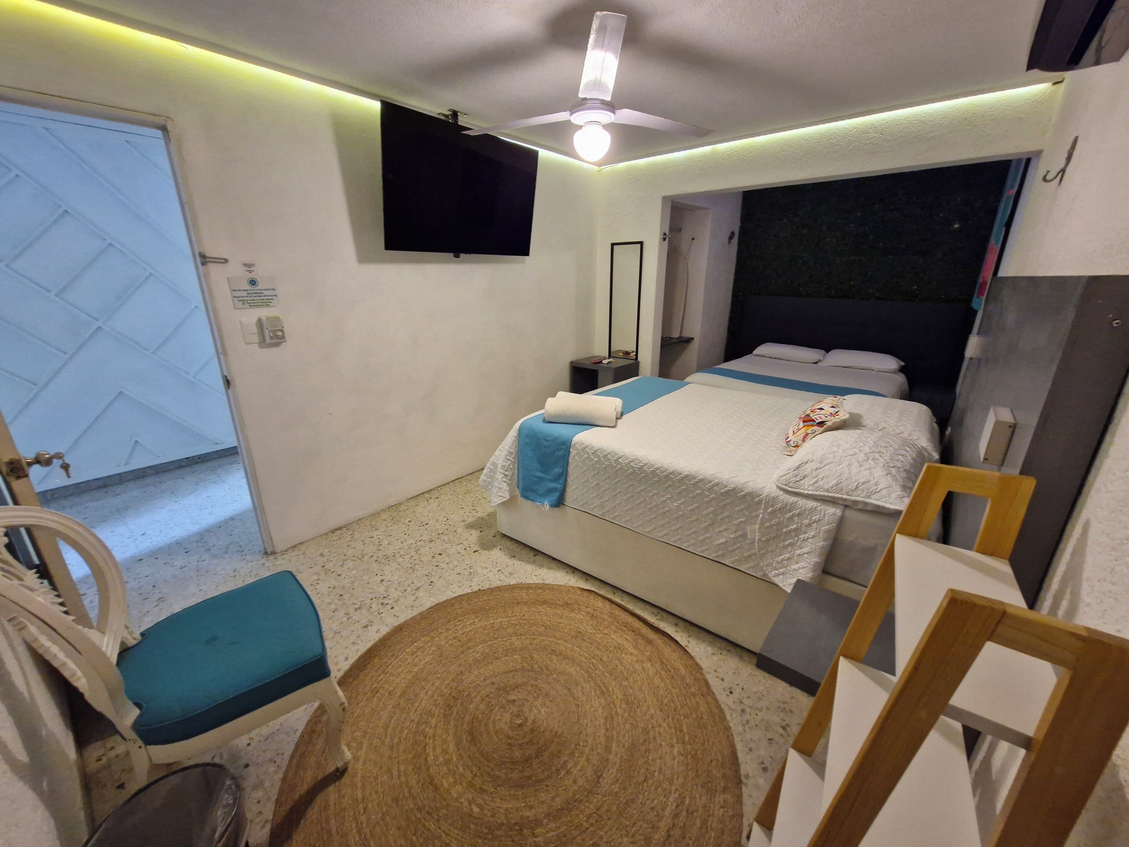 A bedroom with a bed, a chair, a small table, a ceiling fan, and a TV mounted on the wall.
