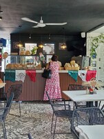 Ένα καφέ με εσωτερικό χώρο, όπου ο πάγκος παρουσιάζει διάφορα γλυκά, υπάρχει ένας ανεμιστήρας οροφής και το δάπεδο έχει διακοσμητικό σχέδιο.