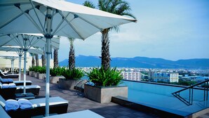 Un salon sur le toit, doté de chaises longues, de parasols et d’une piscine, offrant une vue imprenable sur la ville et les montagnes.