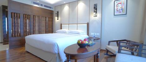 A hotel room with a large bed, a wooden wardrobe, a small table with fruit, and a chair.