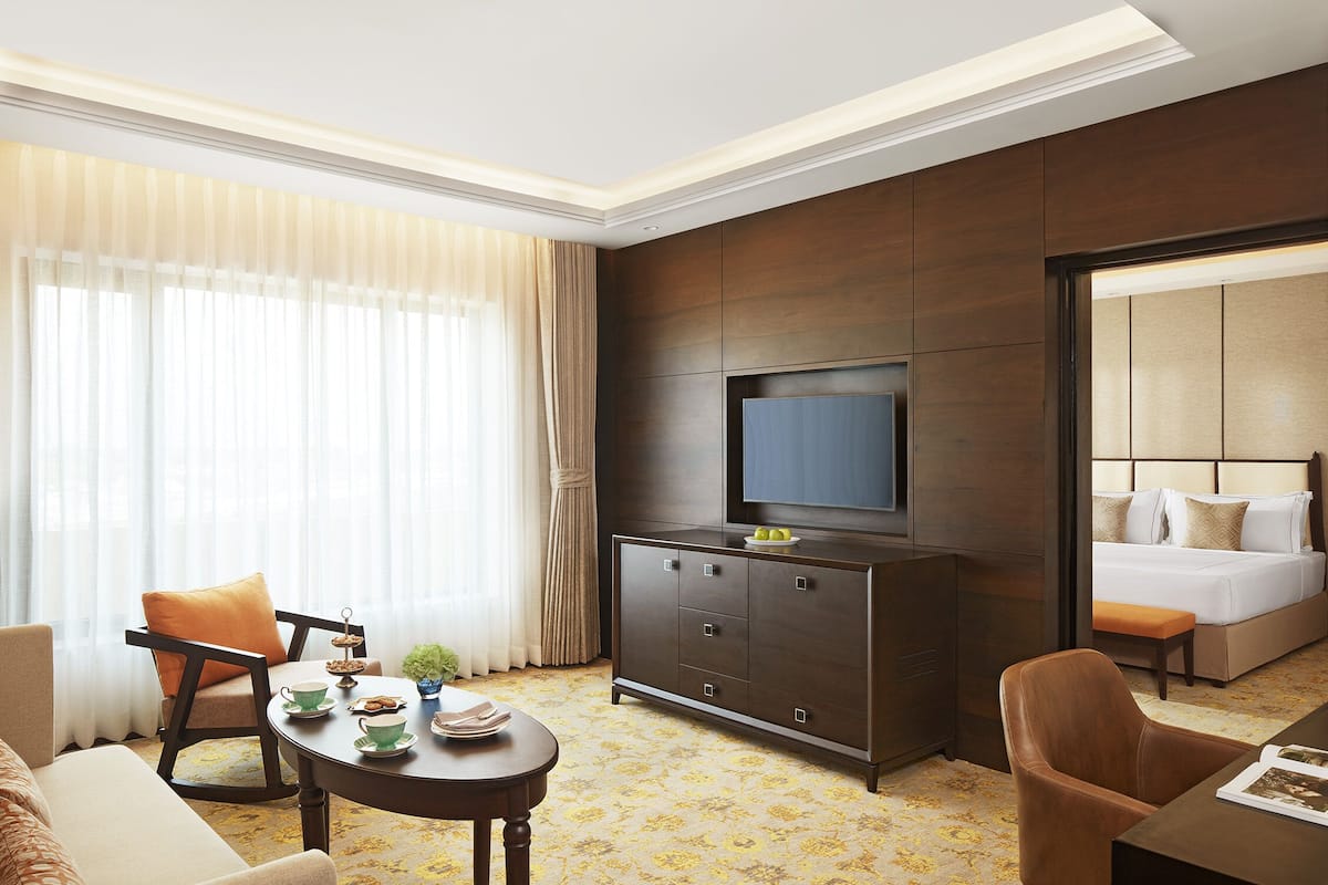A hotel room with a bed, a TV on a cabinet, a dining table with chairs, and a large window with curtains.
