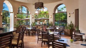 Un espace extérieur aménagé pour les repas, avec plusieurs tables et chaises, de grands parasols et des plantes en pot.