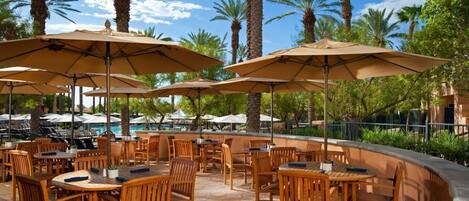An outdoor dining area with wooden tables and chairs, surrounded by palm trees and umbrellas.