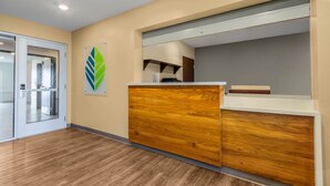 A reception area with a wooden reception desk, a glass door leading to a room, and a wall-mounted green leaf decoration.