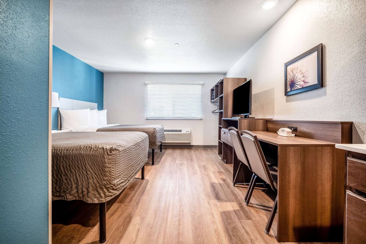 A compact hotel room with two beds, a desk, a chair, a TV, and a window with blinds.