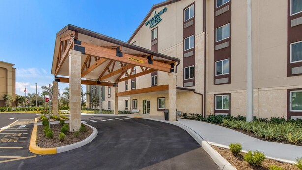 A hotel entrance with a covered walkway, a clear sky, and a landscaped area with small plants. - WoodSpring Suites Tamarac (Tamarac)