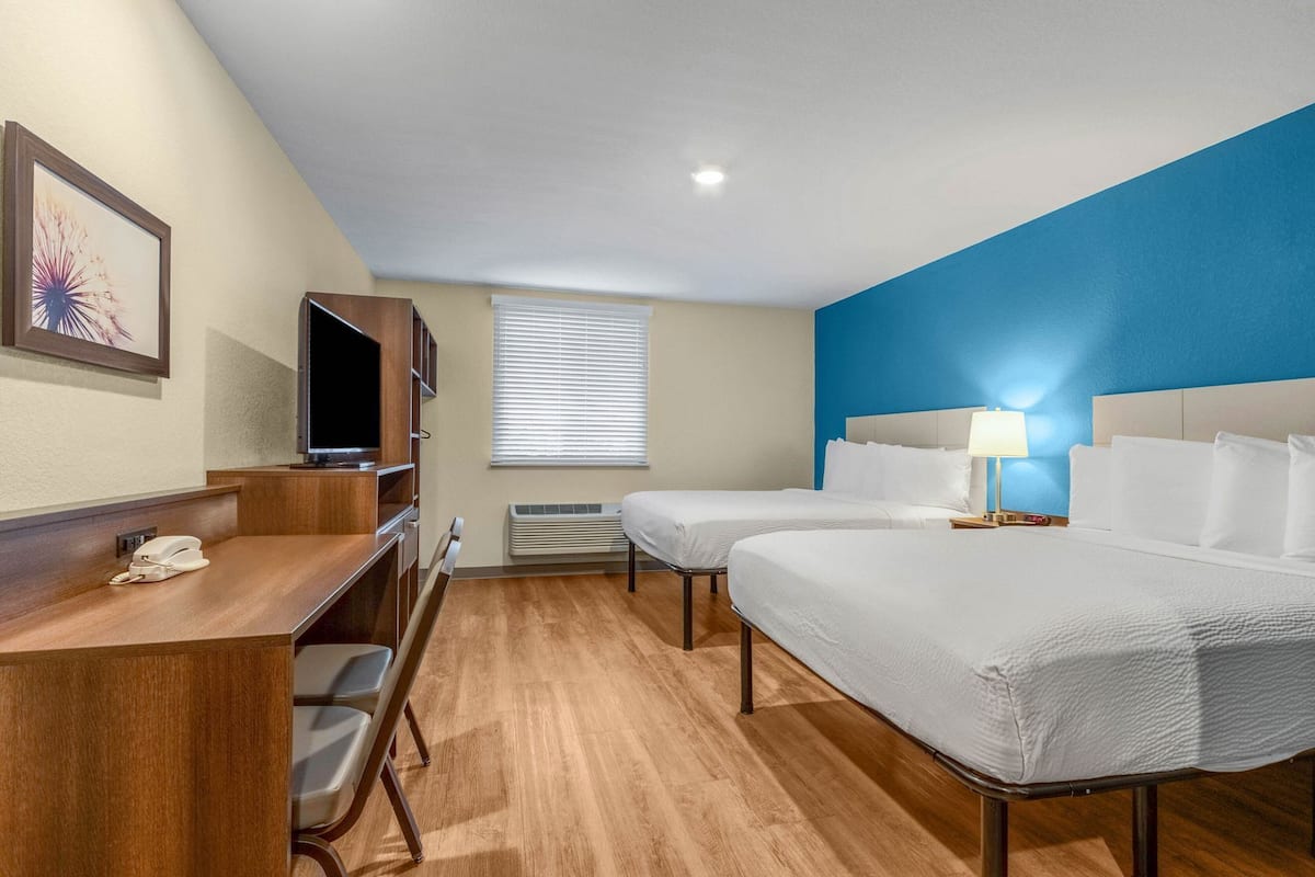 A hotel room with a desk, two beds, a TV, a phone, and a window with blinds.