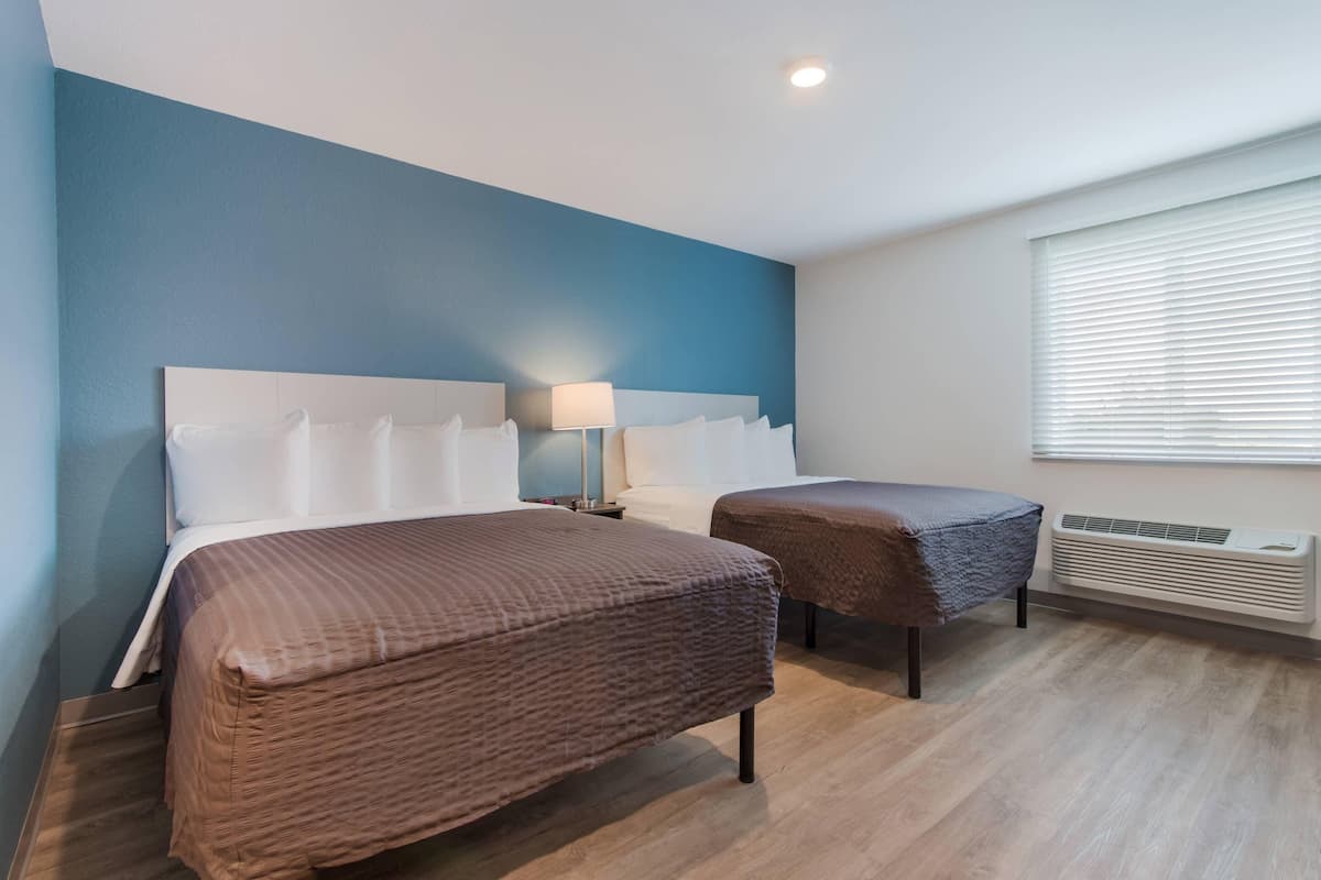 A hotel room with two beds, a nightstand, a lamp, and a window with blinds.