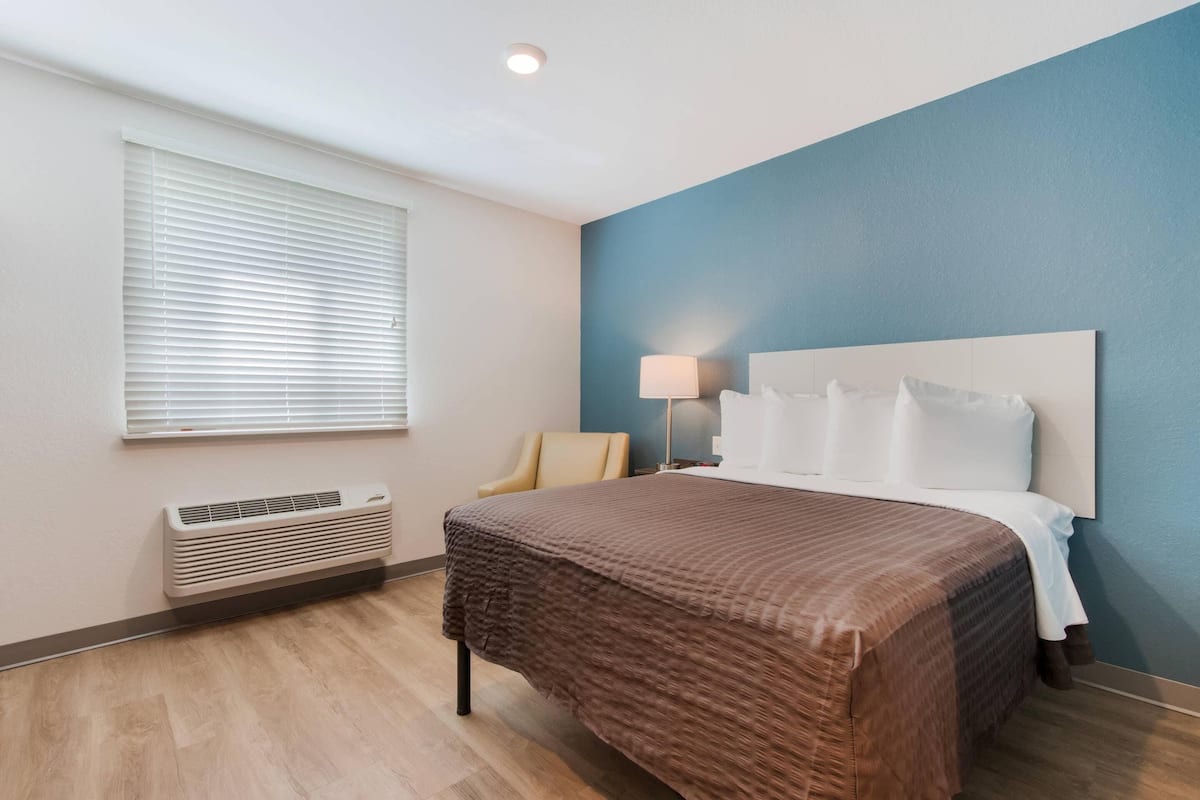 A hotel room with a bed, a chair, a desk, and a window with blinds.