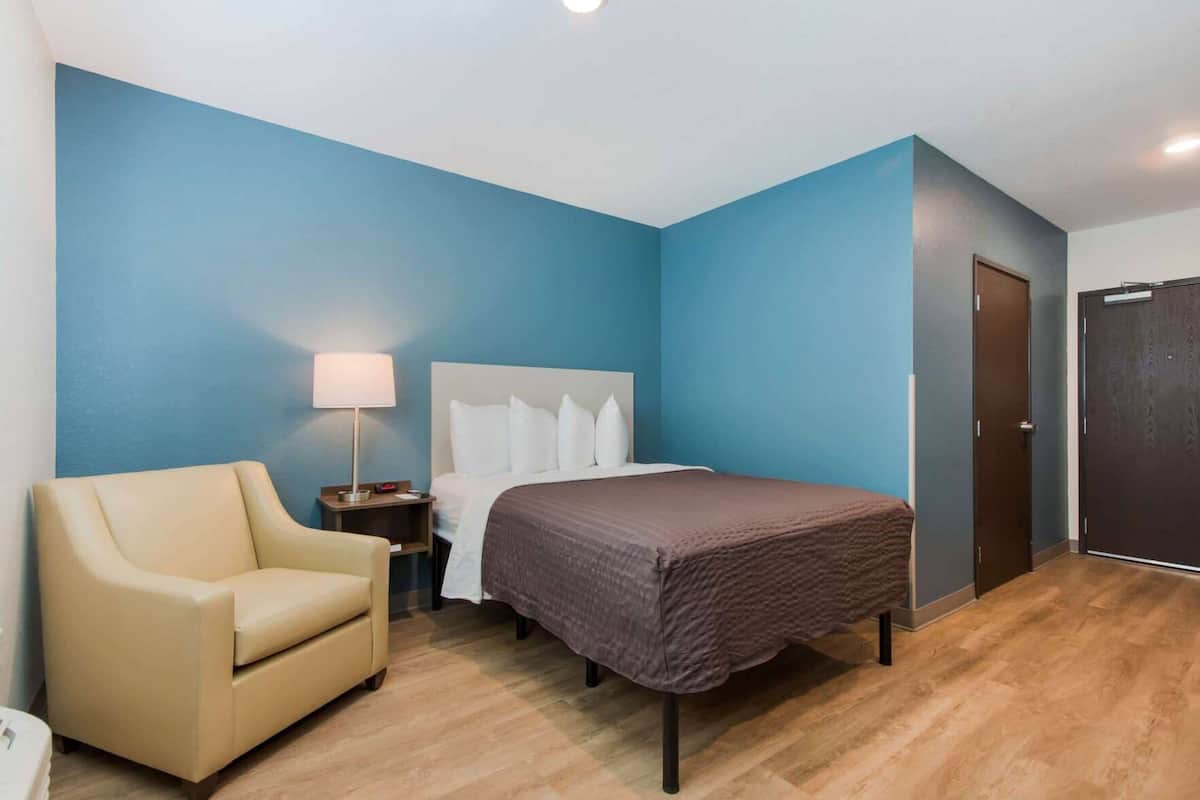 A hotel room with a bed, a chair, a nightstand, and a door.