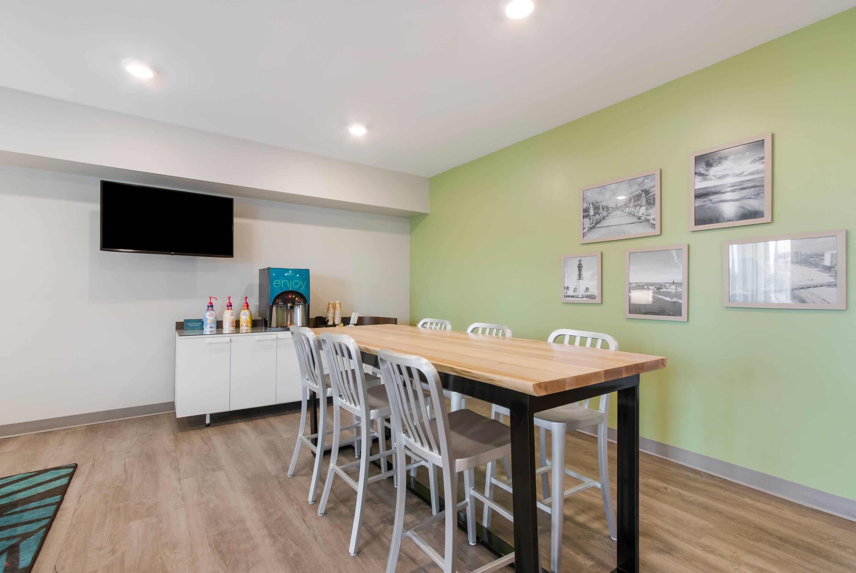 A dining area with a long wooden table, white chairs, a flat-screen TV mounted on the wall, and a small kitchenette with a coffee machine and condiment bottles.
