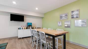 A dining area with a long wooden table, white chairs, a flat-screen TV mounted on the wall, and a small kitchenette with a coffee machine and condiment bottles.