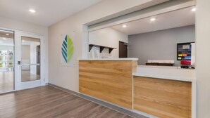 A reception area with a wooden reception desk, a glass door leading to a balcony, and a wall-mounted digital display. - WoodSpring Suites Tamarac (Tamarac)