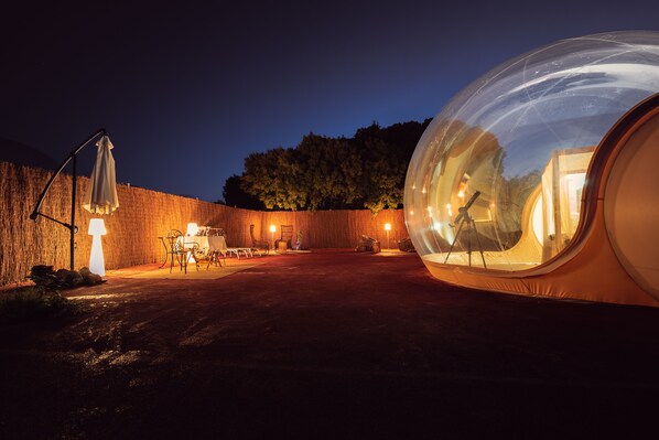 Estructura abovedada transparente que muestra iluminación y mobiliario interior, rodeada por un muro de ladrillo y una valla de madera.