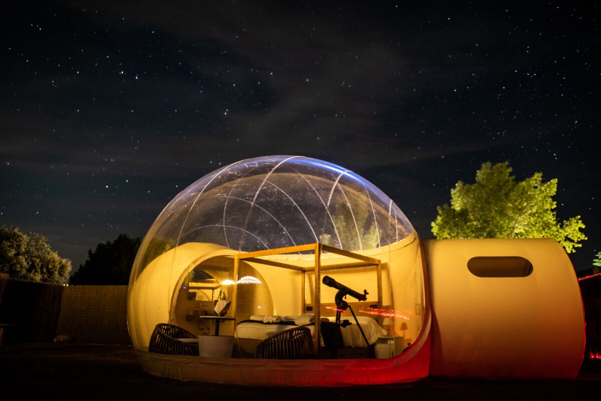 A transparent dome structure with a bed and a telescope inside, set against a night sky.